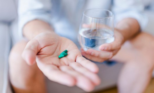 A person holding two tablets in one hand and a glass of water in the other hand.