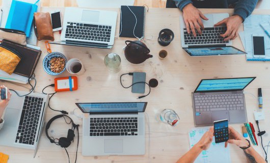 Seen from above, five people are using laptops, with mobiles, drinks and notebooks on the table.