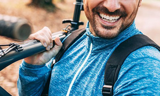 Close up of a smiling man wearing a helmet and carrying a bike over his shoulder.