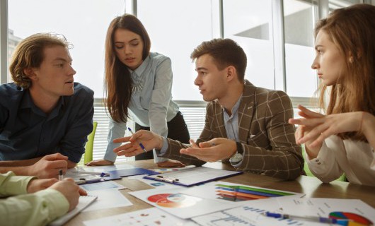 Five smartly dressed people are having a discussion around a table topped with charts and documents.