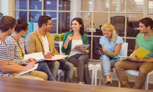 Six men and women sit in a semi-circle in an office setting, holding notepads and discussing ideas.