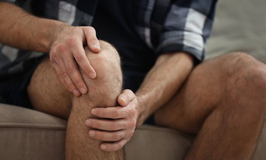 A man is sat on his sofa and holding his knee in pain.