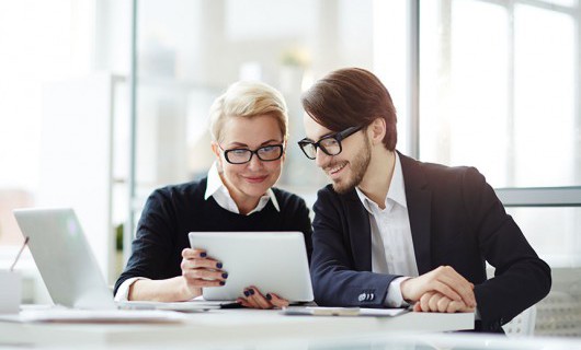 Two smartly dressed professionals smile at a tablet being held by one of the colleagues.