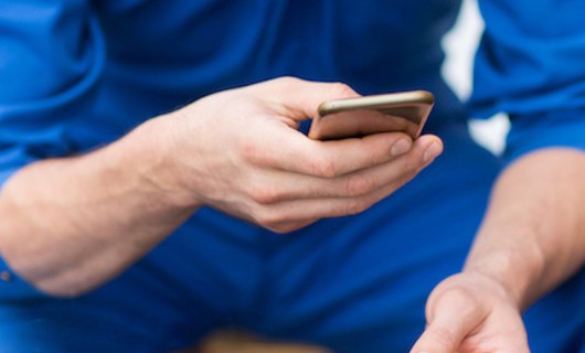 A man in blue overalls is sat looking at his phone and holding a lit cigarette.