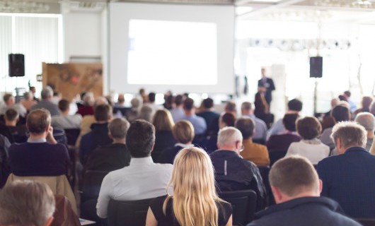 A conference room full of people looks towards a speaker giving a presentation.