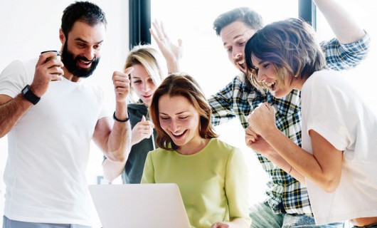 A woman smiles at her laptop whilst a group of people around her cheer happily.