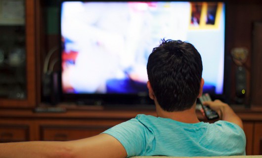 A man is sat holding a television remote, looking and pointing the remote towards the television.