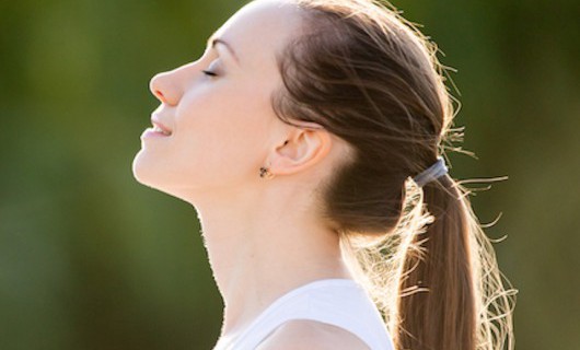 A young woman stands with her eyes closed, smiling and looking up at the sun, looking peaceful.