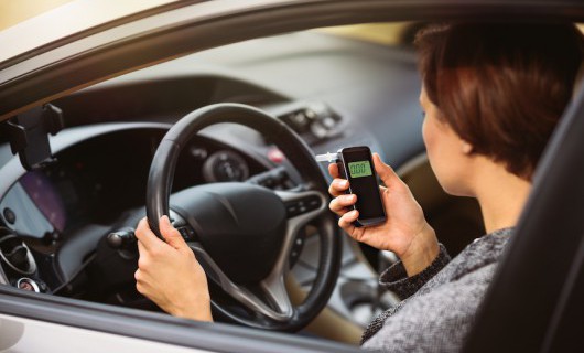 A woman is sat in the driver seat in her car, looking down at the breathalyser device in her hand.