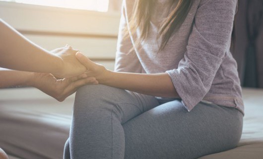 A woman, sat on the edge of a bed, holds hands with another person in front of her.